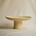 A minimalist ceramic pedestal plate in a warm sandy beige color, set against a softly draped fabric backdrop.